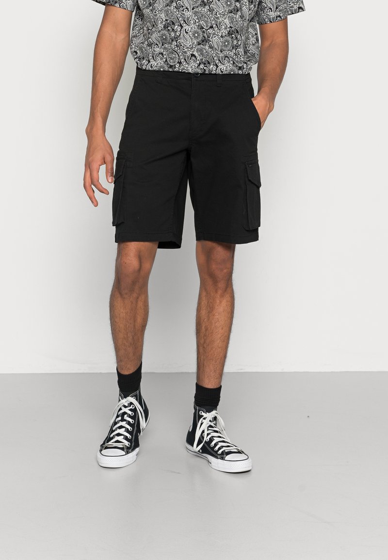 Homme portant un short cargo noir, des baskets montantes noires avec des lacets blancs, des chaussettes noires et une chemise à manches courtes à motifs.