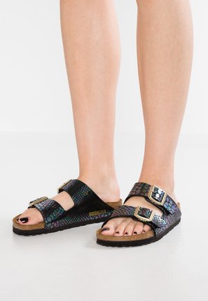 Pieds portant des sandales Birkenstock noires iridescentes avec deux brides à boucle et des semelles en liège sur un sol blanc.