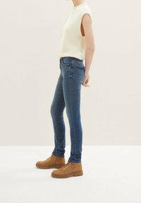 Úzké modré džíny s vysokým pasem a pěti kapsami, kombinované s bílým bezramenným pleteným topem a hnědými kotníkovými botami.