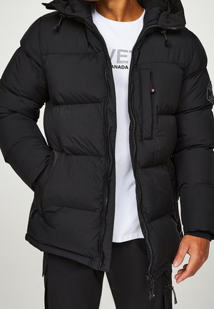 Mężczyzna w czarnej, rozpiętej, pikowanej zimowej kurtce na białym T-shircie z logo i czarnych spodniach, z rękami opuszczonymi wzdłuż ciała, na jasnym tle.