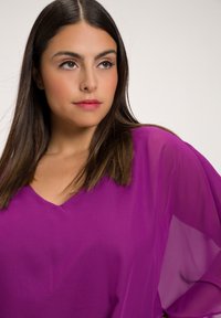 Violetti, läpinäkyvä paita, jossa on V-muotoinen kaula-aukko ja valuvaa hihaa. Kangas näyttää kevyeltä, sillä on sileä tekstuuri ja kirkas väri.