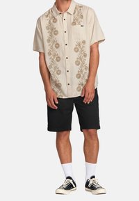 Camisa de manga corta beige con bordado floral marrón, diseño de botones, bolsillo en el pecho izquierdo; combinada con pantalones cortos negros y zapatillas negras.