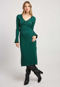 Groene geribde lange jurk met een V-hals en uitlopende mouwen, met een getailleerde pasvorm en een lengte tot onder de knie, gecombineerd met zwarte kniehoge laarzen.