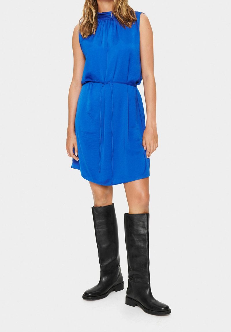 Abito blu senza maniche con scollo arricciato e vita con coulisse, abbinato a stivali neri in pelle al ginocchio. Tessuto liscio, design casual.