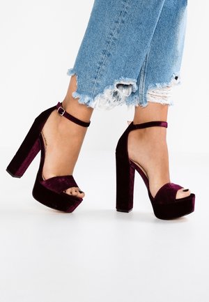 Fødder iført bordeauxrøde fløjl platform hæle med ankel stropper, kombineret med lyseblå slidte jeans med frynsete kanter.