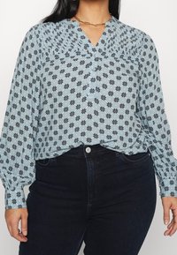 Jasnoniebieska bluzka z czarnym kwiatowym wzorem, zapinana na guziki z przodu, z plisowanym dekoltem i długimi rękawami, połączona z ciemnymi spodniami dżinsowymi o wysokim stanie.