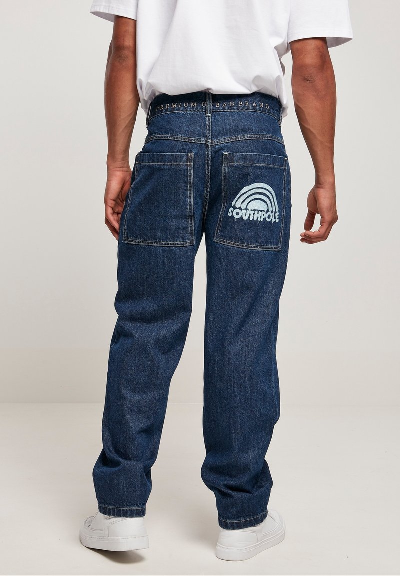 Personne portant un jean denim bleu foncé avec de grandes poches arrière, un texte blanc et un motif arc-en-ciel sur la poche droite, des baskets blanches et une chemise blanche.