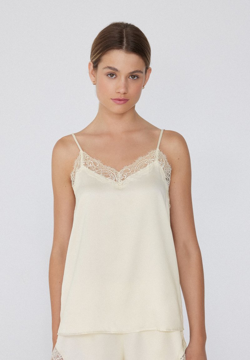 Camisole en satin couleur crème avec des détails en dentelle délicate le long du décolleté et des bretelles. Texture douce, coupe droite, bretelles spaghetti réglables.
