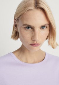 Lys lilla T-shirt med rund hals og korte ærmer, med en glat stoffer tekstur. Modellen har kort blond hår og klare blå øjne.