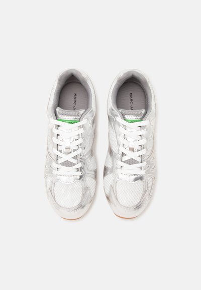 Paire de baskets blanches et argentées avec des lacets blancs et une étiquette de marque verte sur la languette, vue de dessus sur fond blanc.