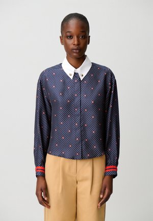 Mujer con una blusa azul marino con estampado, cuello blanco y pantalones beige, de pie frente a un fondo claro liso.