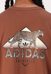Brūns kokvilnas T-krekls ar lielu leoparda grafiku, uz kura ir uzraksts "ADIDAS" bieza, leoparda raksta burtos uz gaiša fona.