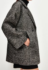 Strukturierter Mantel im Fischgrätenmuster in Schwarz und Weiß. Verfügt über ein doppelreihiges Design, große Revers und Seitentaschen.