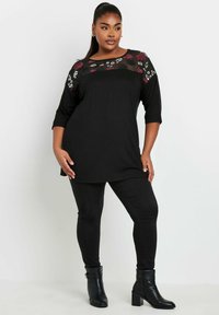 Mustat tunikatoppi, jossa on kukkakirjontaa läpikuultavalla paneelilla, yhdistetty mustiin tiukkoihin housuihin ja nilkkabootseihin. Pehmeä kangas ja rento malli.