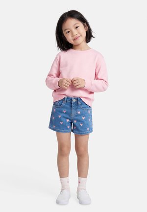 Fiatal lány mosolyog, rózsaszín pulóvert, szív alakú mintás farmer rövidnadrágot, világos rózsaszín zoknit szív mintával és fehér vászoncipőt visel.