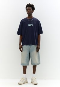 Navy oversized t-shirt with logo print, paired with light blue denim shorts. White socks and beige sneakers complete the outfit.