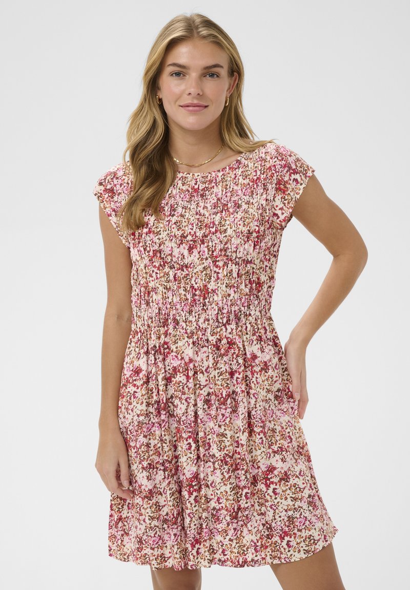 Giovane donna che indossa un vestito floreale a maniche corte e lunghezza al ginocchio, nei toni del rosa, rosso e beige, in piedi con una mano sul fianco.