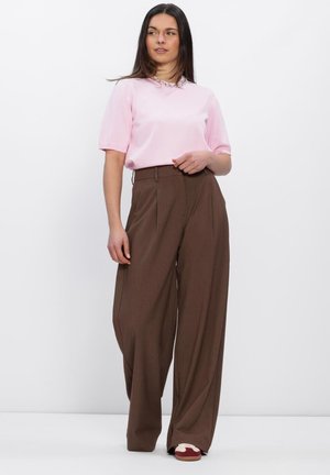 Vrouw staat getooid met een lichtroze top met korte mouwen en versierde kraag, wijde bruine broek en rood-witte sneakers tegen een witte achtergrond.