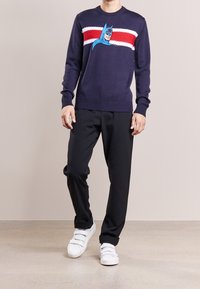 Marineblauwe trui met rode en witte horizontale strepen, voorzien van een geborduurd blauw Batman ontwerp op de voorkant. Gecombineerd met zwarte broeken en witte sneakers.