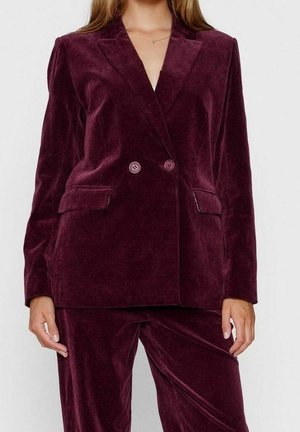 Femme portant un blazer en velours côtelé violet foncé à double boutonnage et un pantalon assorti, debout avec les bras détendus le long du corps.