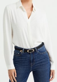Weiße Bluse mit langen Ärmeln und strukturiertem Muster; klassischer Kragen und durchgehende Knopfleiste. Kombiniert mit dunkelblauen Jeans und einem schwarzen Gürtel.