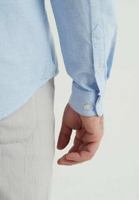 Chemise à manches longues bleu clair avec des poignets boutonnés, en tissu texturé et coupe ajustée, portée avec un pantalon de couleur claire.