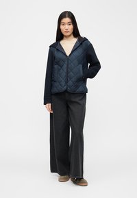 Jeune femme portant une veste matelassée bleu marine zippée, une chemise beige, un jean noir à jambe large, et des baskets marron, debout devant un fond blanc.