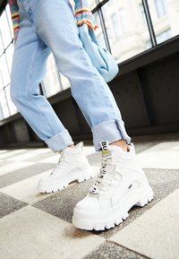 Sneaker bianche chunky con suola testurizzata, design con lacci e dettagli di branding. Indossate con jeans azzurri a risvolto in un ambiente interno luminoso.