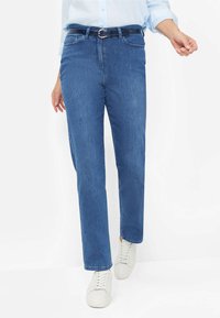 Jeans in denim blu con taglio dritto, dotati di una cintura scura, tasche frontali e una texture liscia, abbinati a sneakers bianche.
