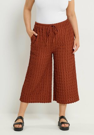 Kvinde iført rustfarvede, teksturerede, vidde-lårkortbukser med snøre i taljen, hvid top og sorte platformsandaler, stående med hænderne i lommerne.