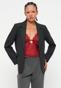 Blazer noir à carreaux avec revers crantés, deux poches avant et fermeture à un bouton, associé à un haut rouge ornée de dentelle et à un pantalon gris.