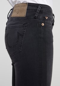 Jeans de mezclilla negra con un parche de cuero de True Religion, bolsillos traseros decorados con un diseño de pedrería y acentos de hardware en color cobre.