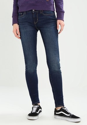 Personne portant un jean skinny bleu foncé, des baskets noires et blanches, et un sweat-shirt violet, les mains détendues le long du corps.