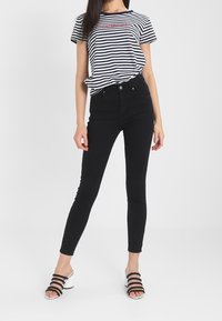 Camiseta de rayas navy y blanca con un logo gráfico, combinada con unos jeans negros ajustados de cintura alta y sandalias de tacón con acentos plateados.