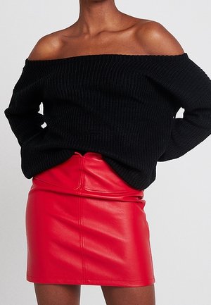 Zwart geribbelde off-shoulder trui gecombineerd met een hooggetailleerde, rode leren minirok op een staand persoon.