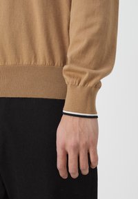 Gros plan sur la main gauche d'une personne, détendue à côté d'un pantalon noir, portant un pull camel avec des poignets côtelés arborant de fines rayures noires et blanches.