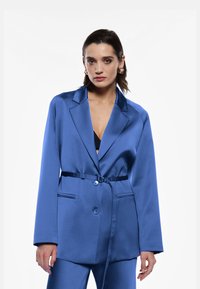 Blazer satin blu con rever a punta, chiusura con un solo bottone, tasche laterali e cintura coordinata. Texture liscia ed design elegante.