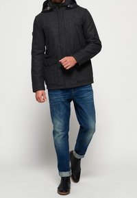 Donkergrijze hooded jas gemaakt van een wolmix, met zijzakken en een geruite voering. Gecombineerd met een blauwe spijkerbroek en zwarte leren laarzen.