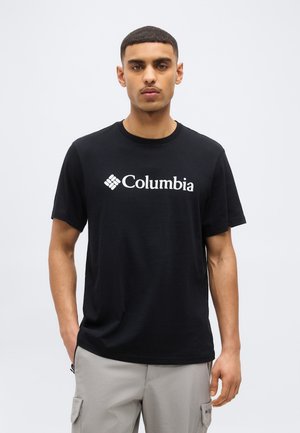 Giovane uomo in piedi con le mani nelle tasche, indossa una maglietta nera Columbia e pantaloni cargo grigio chiaro su uno sfondo semplice.