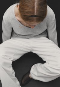 Gray sweater with a wide neckline, paired with loose-fitting white pants. The model is seated with a black background and dark footwear.