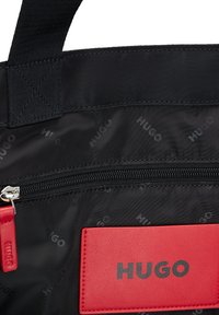 Borsa tote in nylon nera con etichetta in pelle rossa, tasca con zip e logo impresso. Presenta un materiale testurizzato e spallacci robusti.