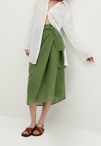 Jupe portefeuille en lin vert avec un grand nœud à l'avant, associée à une chemise blanche boutonnée ample et des sandales marron. Tissu texturé visible.