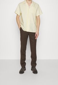 Vaaleankeltainen, lyhythihainen napillinen paita, jossa pystysuorat raidat, yhdistetty ruskeisiin housuihin ja tummiin kenkiin. Yksinkertainen pinta ja rintatasku.