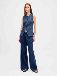 Woman standing confidently wearing sleeveless buttoned dark denim vest and wide-leg jeans with black open-toe sandals on white background.