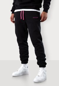 Schwarze Jogginghose mit pinkem Kordelzug und Logo. Verfügt über elastische Bündchen und einen lockeren Schnitt. Kombiniert mit weißen Sneakers und Socken.