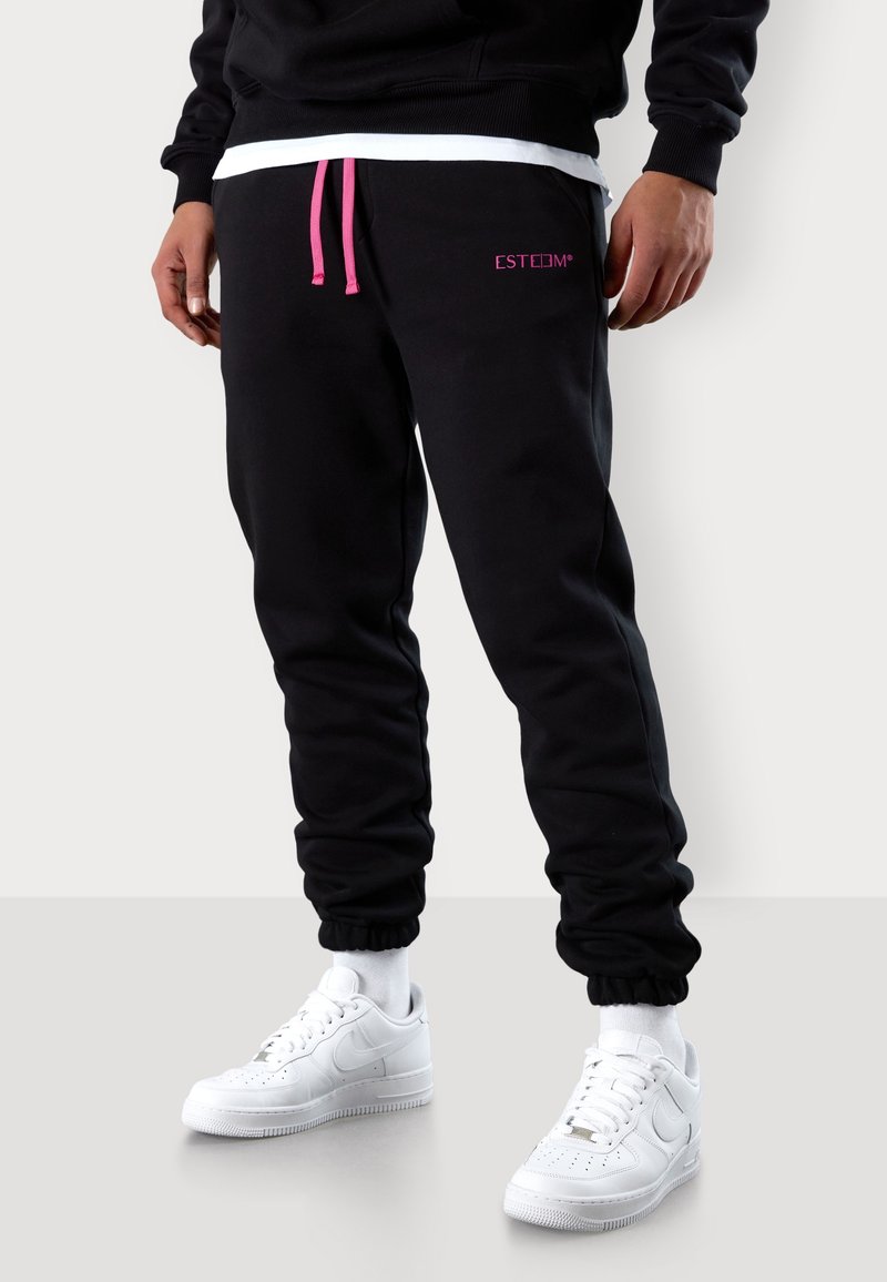 Schwarze Jogginghose mit pinkem Kordelzug und Logo. Verfügt über elastische Bündchen und einen lockeren Schnitt. Kombiniert mit weißen Sneakers und Socken.