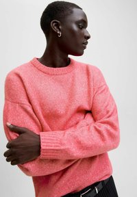 Persoon met kort haar in een koralen roze sweater, met gekruiste armen, die naar de zijkant kijkt tegen een effen lichte achtergrond.