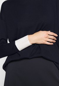 Personne portant un haut noir à manches longues avec un poignet côtelé blanc, main reposant sur le côté du torse, pantalon noir visible en dessous.