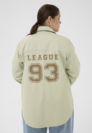 Femme aux cheveux foncés attachés en queue de cheval basse porte une chemise oversize vert clair avec l'inscription "LEAGUE 93" en marron dans le dos.