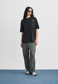 Μαύρο oversize t-shirt, γκρι παντελόνι cargo με πολλές τσέπες και γκρι αθλητικά παπούτσια. Το μοντέλο φοράει γυαλιά ηλίου και στέκεται σε μπλε χαλί.